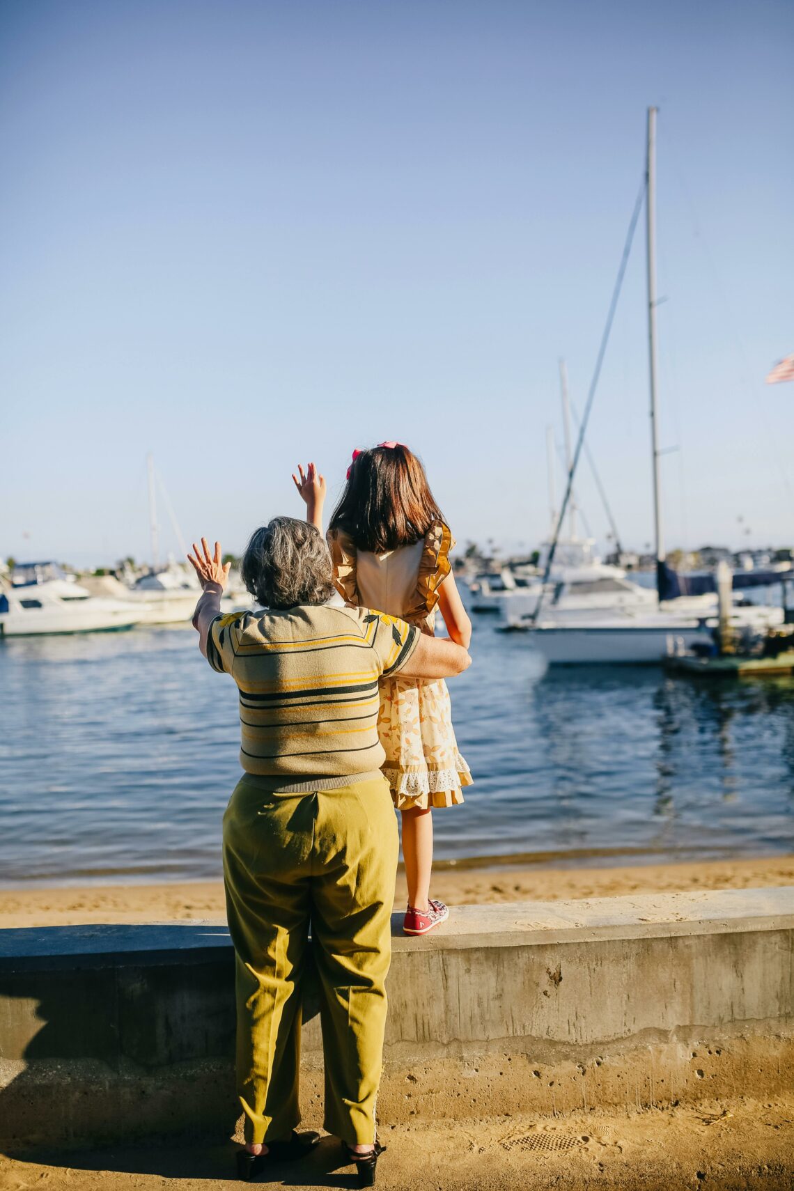 An older woman standing beside a young girl at a waterfront, both facing the water and waving toward boats in the marina on a bright, sunny day.