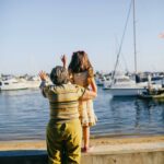 An older woman standing beside a young girl at a waterfront, both facing the water and waving toward boats in the marina on a bright, sunny day.