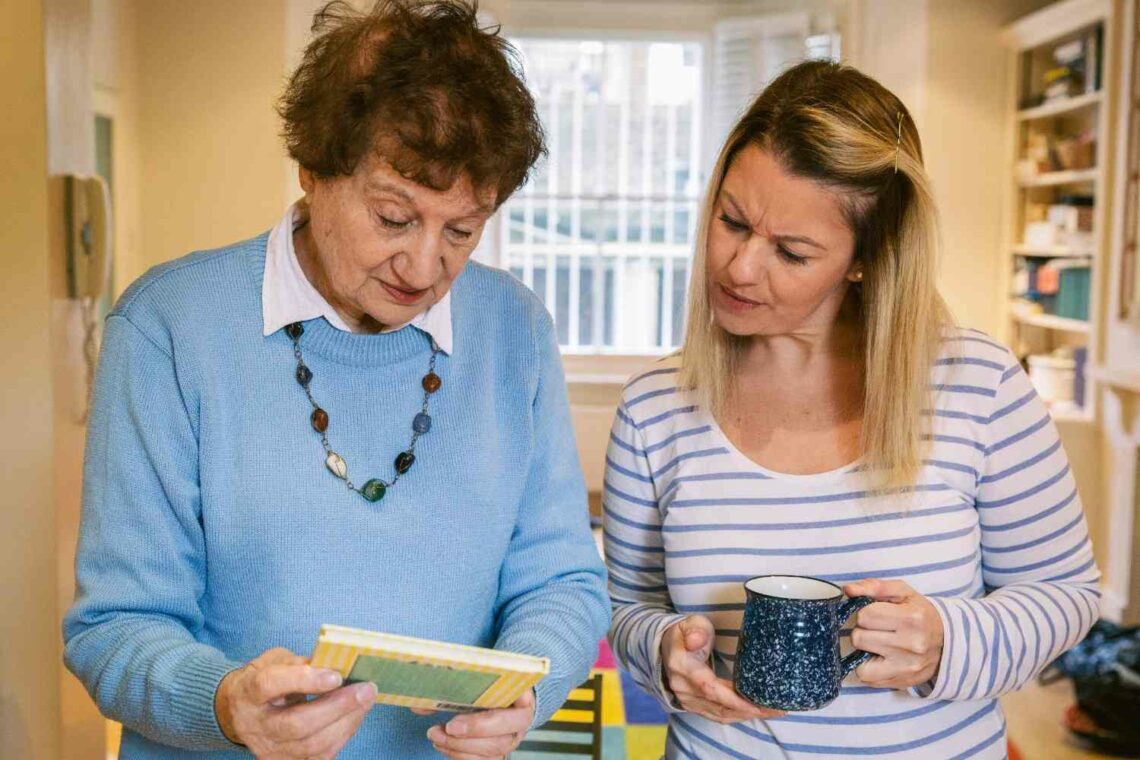 An older woman and a younger woman stand together in a home, looking at a piece of paper while talking, with the younger woman holding a mug.