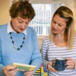 An older woman and a younger woman stand together in a home, looking at a piece of paper while talking, with the younger woman holding a mug.