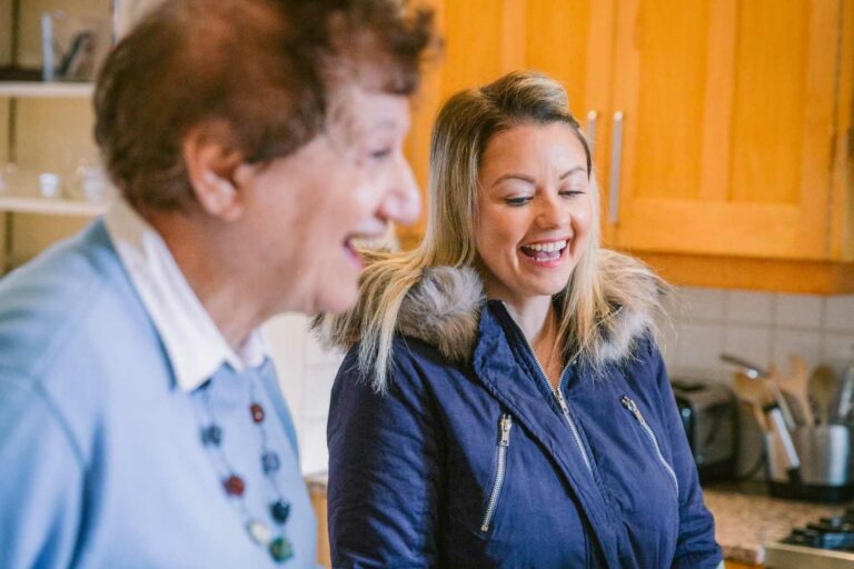 A younger woman and an older woman smiling and chatting together in a warm, home kitchen setting