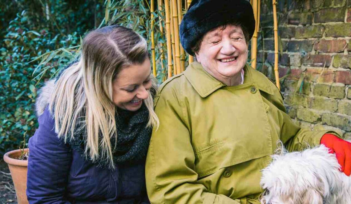Older woman in a green coat and red gloves smiling while sitting outside with a younger woman and gently petting a fluffy white dog.