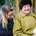 Older woman in a green coat and red gloves smiling while sitting outside with a younger woman and gently petting a fluffy white dog.