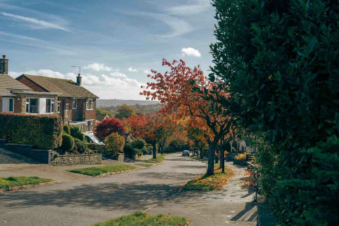 A quiet residential street lined with detached houses and autumn trees in shades of red and orange, with rolling hills visible in the background.