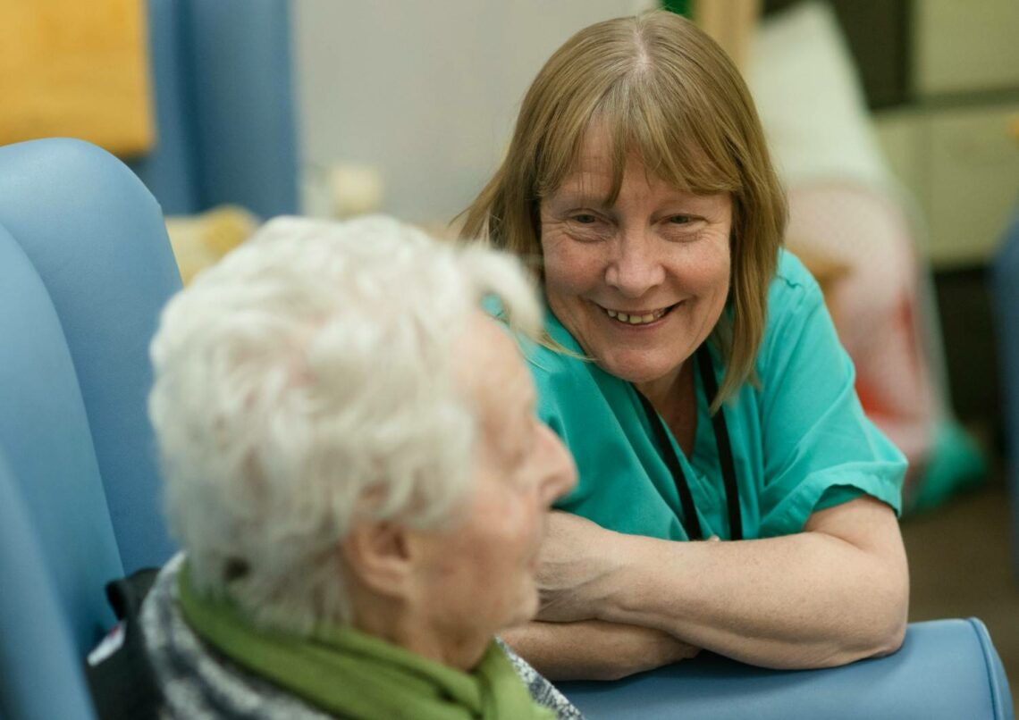 A carer in teal scrubs leaning forward to speak with an older woman seated in a blue chair