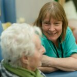A carer in teal scrubs leaning forward to speak with an older woman seated in a blue chair