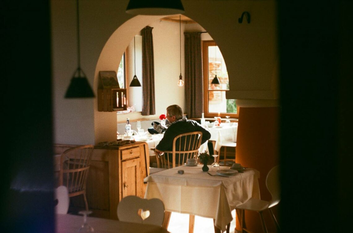 Older man seated alone at a dining table in a warmly lit room with wooden furniture and an arched doorway