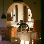 Older man seated alone at a dining table in a warmly lit room with wooden furniture and an arched doorway