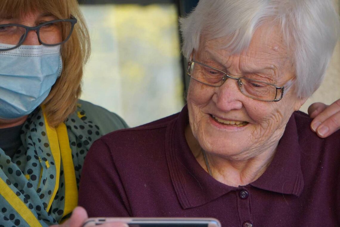 An older woman with white hair and glasses looking at a phone screen, sitting next to a masked caregiver in a patterned top