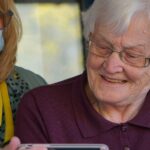An older woman with white hair and glasses looking at a phone screen, sitting next to a masked caregiver in a patterned top