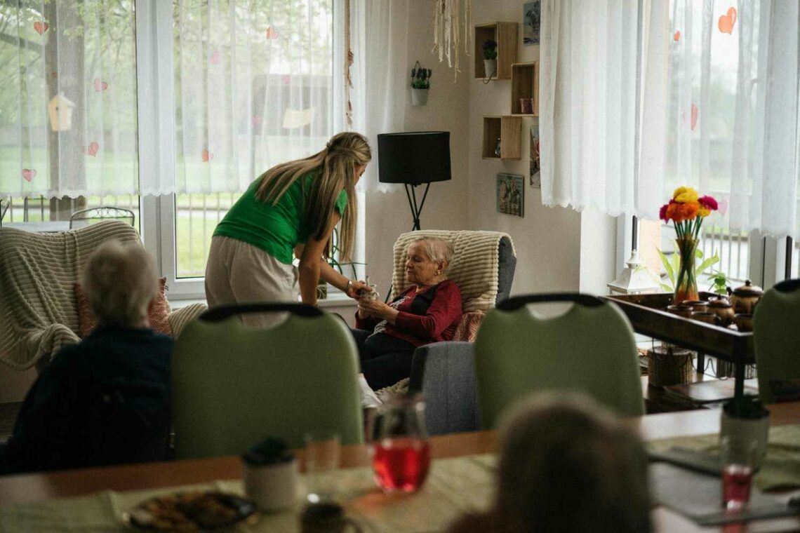 A younger woman leaning towards an older woman seated in an armchair in a bright living room, handing her a small object