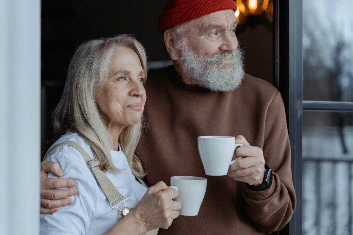 Two older adults standing by a window holding mugs, the man with his arm around the woman's shoulders