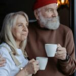 Two older adults standing by a window holding mugs, the man with his arm around the woman's shoulders