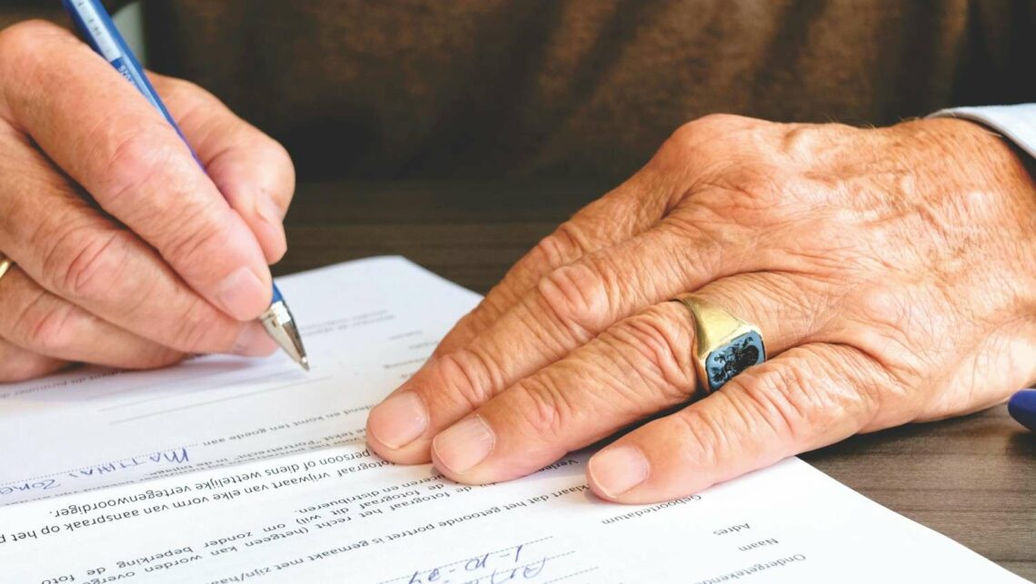 Older man's hands signing a legal document, with a gold ring visible on his right hand