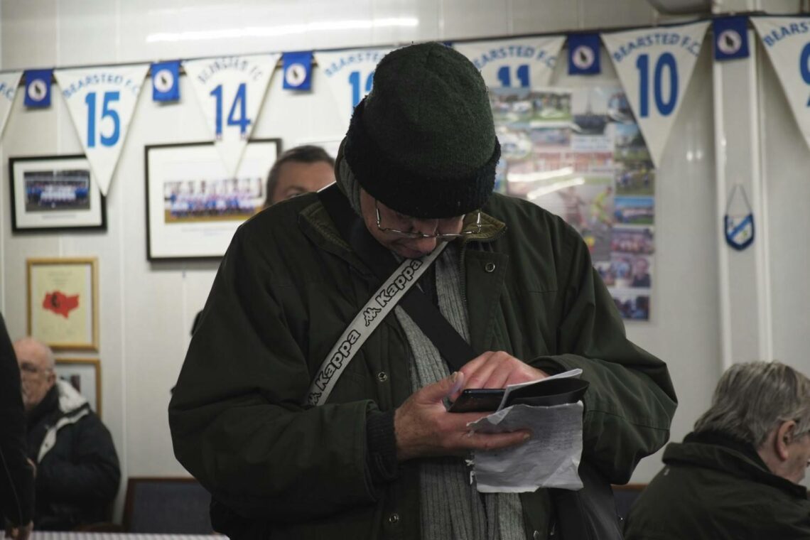 An older man in a green winter hat and jacket looking at his phone and papers inside a football club room with blue and white bunting