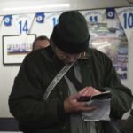 An older man in a green winter hat and jacket looking at his phone and papers inside a football club room with blue and white bunting