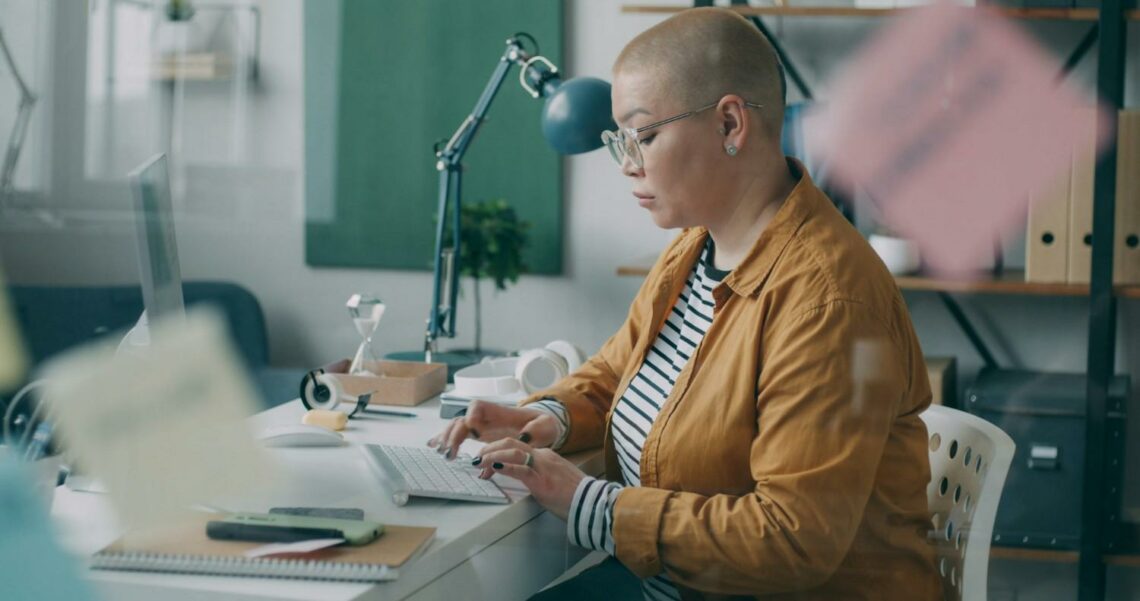 A middle-aged woman with a shaved head sits at a desk working at a computer in a bright office environment.