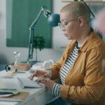 A middle-aged woman with a shaved head sits at a desk working at a computer in a bright office environment.