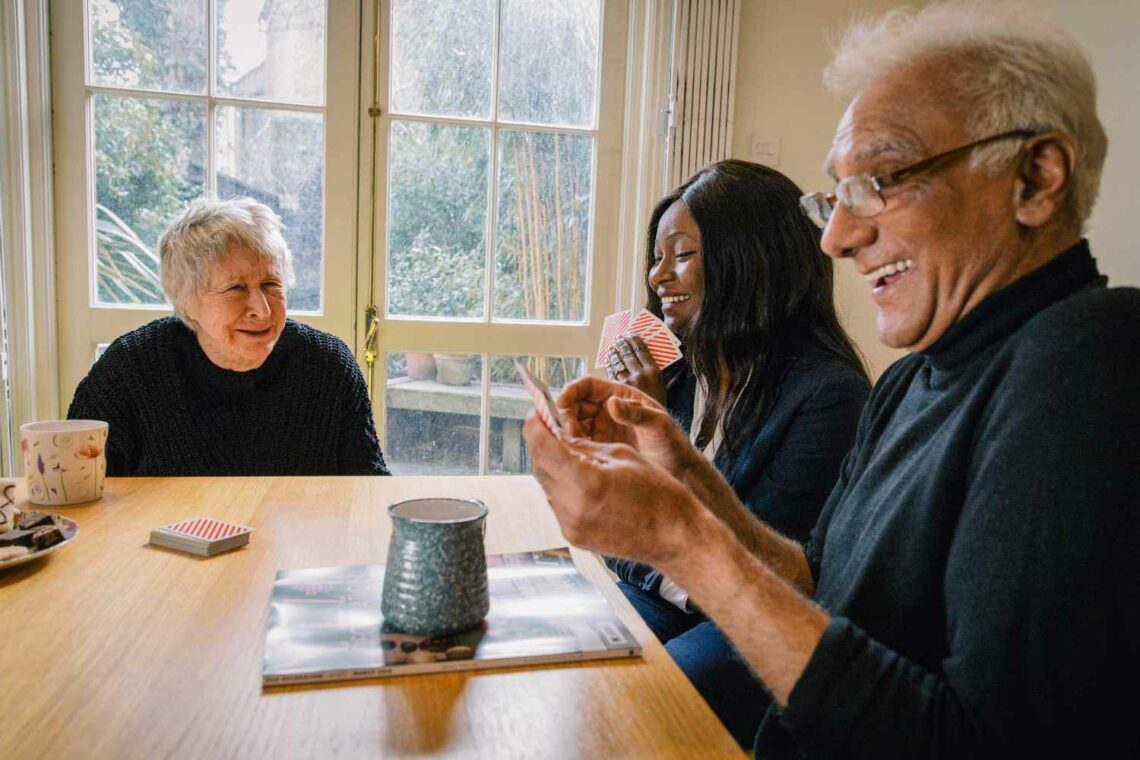 A carer and two older adults playing cards together at a kitchen table at home