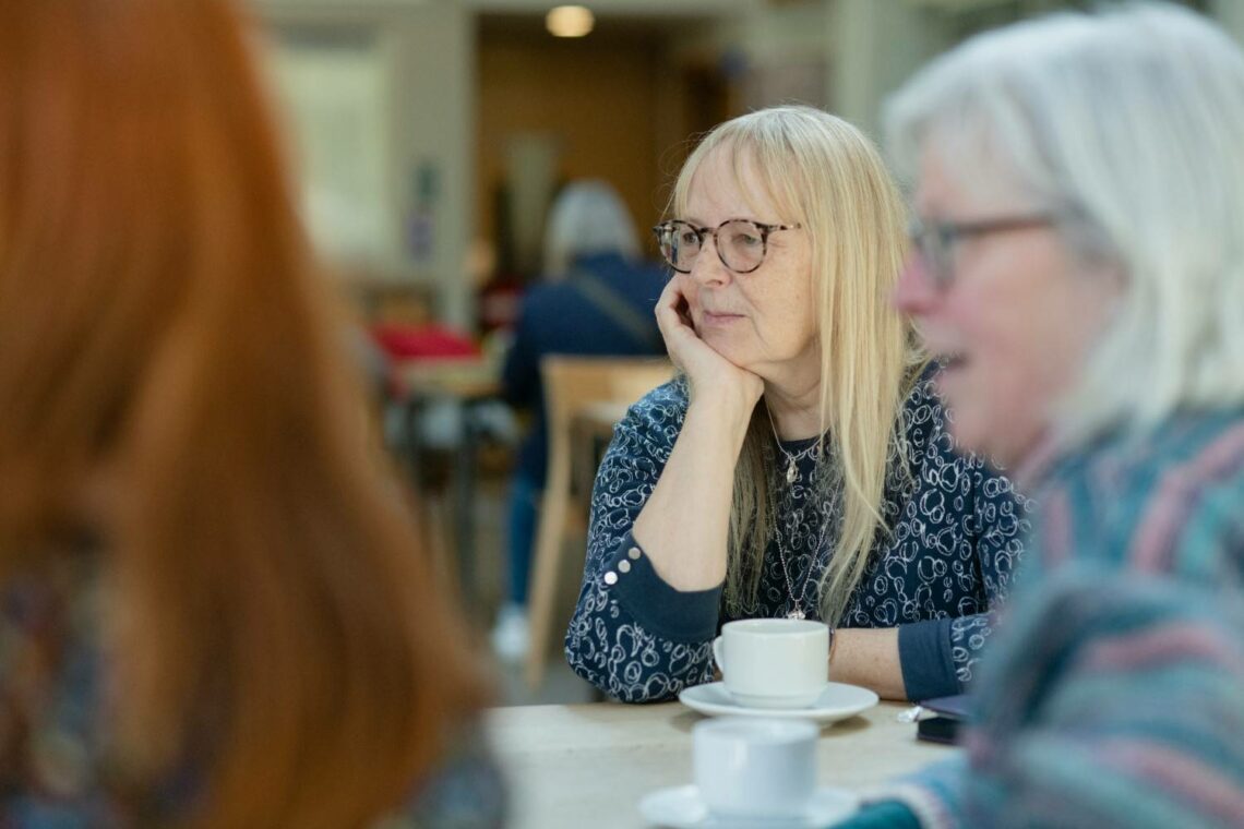 Two women seated at a cafe table in conversation, one with blonde hair resting her chin on her hand, a white cup and saucer in front of her