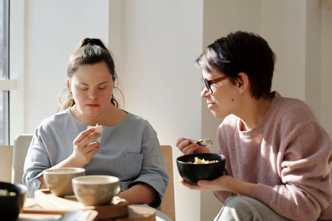 Two women enjoying a meal together in a cozy kitchen, surrounded by cooking utensils and fresh ingredients.
