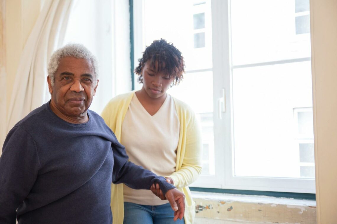 An older man and woman stand together in front of a large window, smiling and enjoying each other's company.