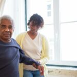 An older man and woman stand together in front of a large window, smiling and enjoying each other's company.