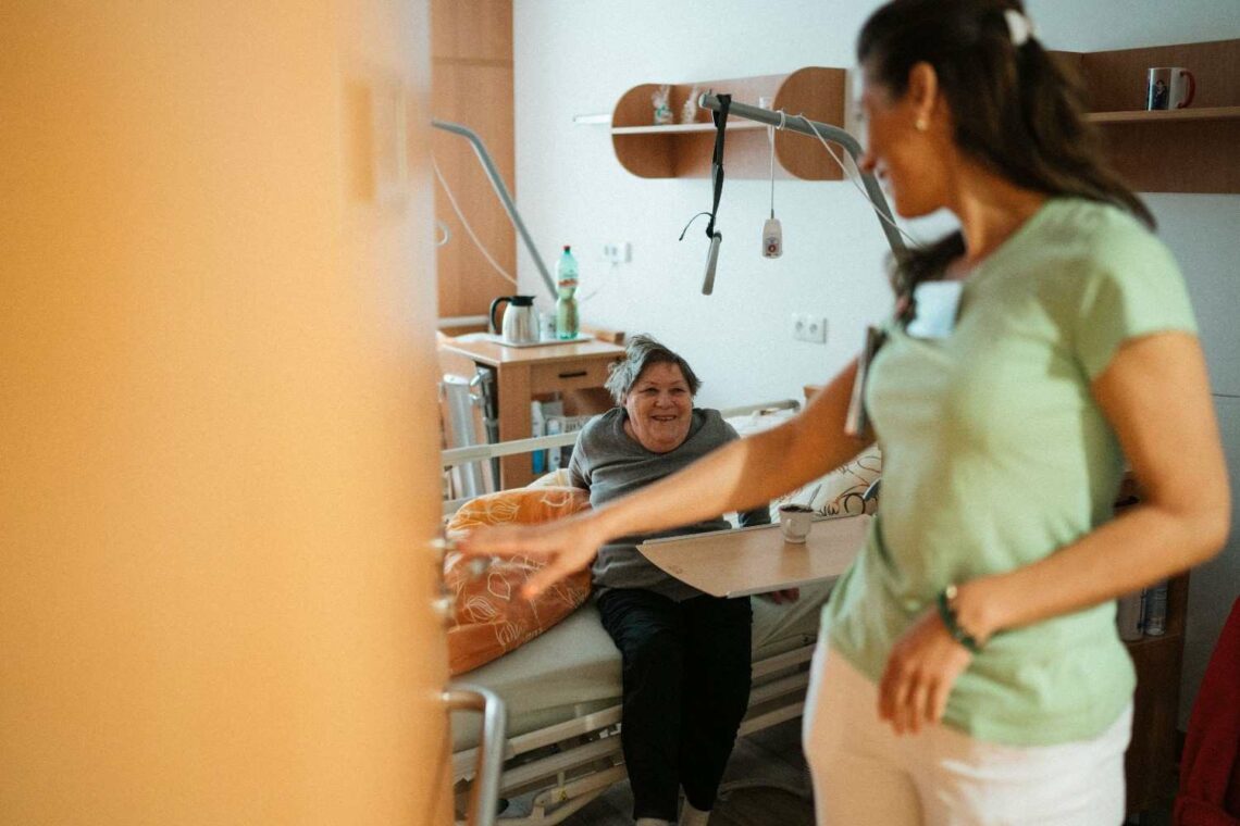 A woman in scrubs converses with a patient in a hospital room, providing care and support.