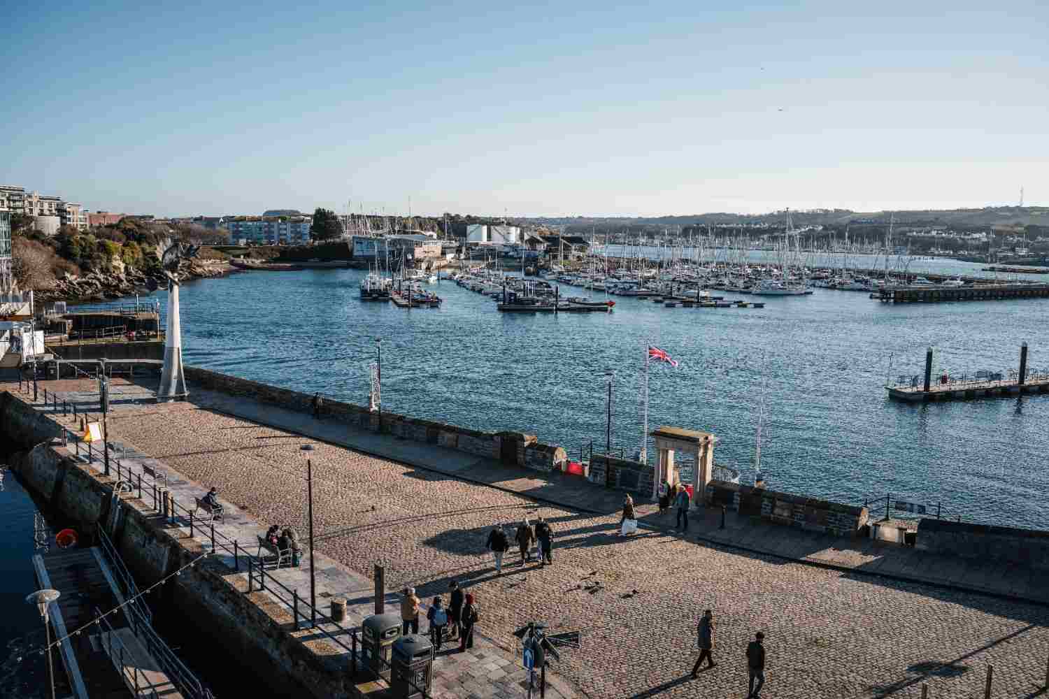 People stroll along a pier with boats docked in a marina, enjoying a sunny day by the water.